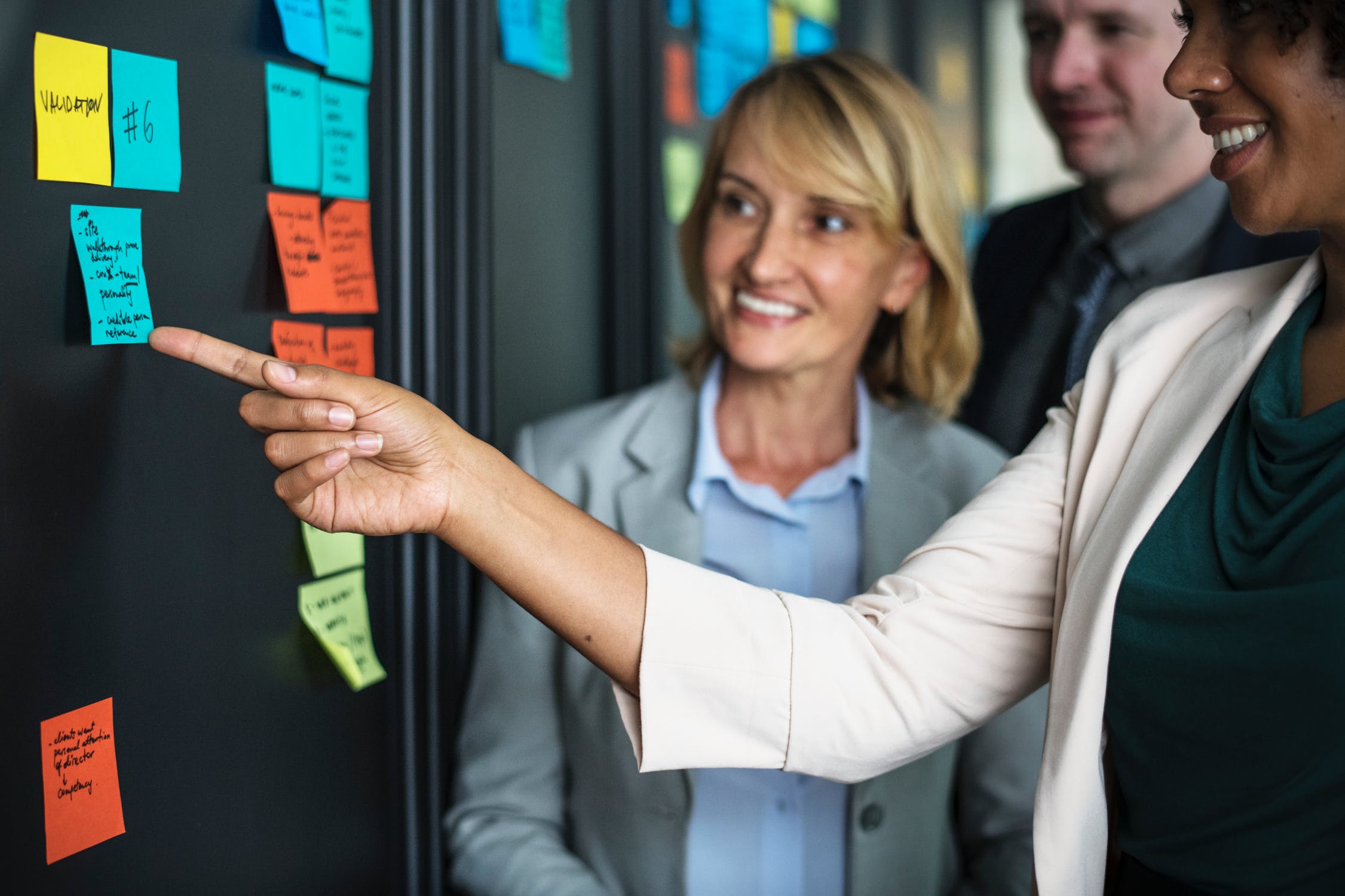 Woman Pointing at Sticky Note on Wall with Coworkers Woman Pointing at Sticky Note on Wall with Coworkers