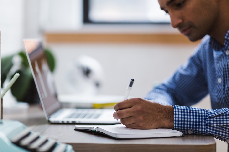 Man writing in notebook on desk in front of a laptop Man writing in notebook on desk in front of a laptop